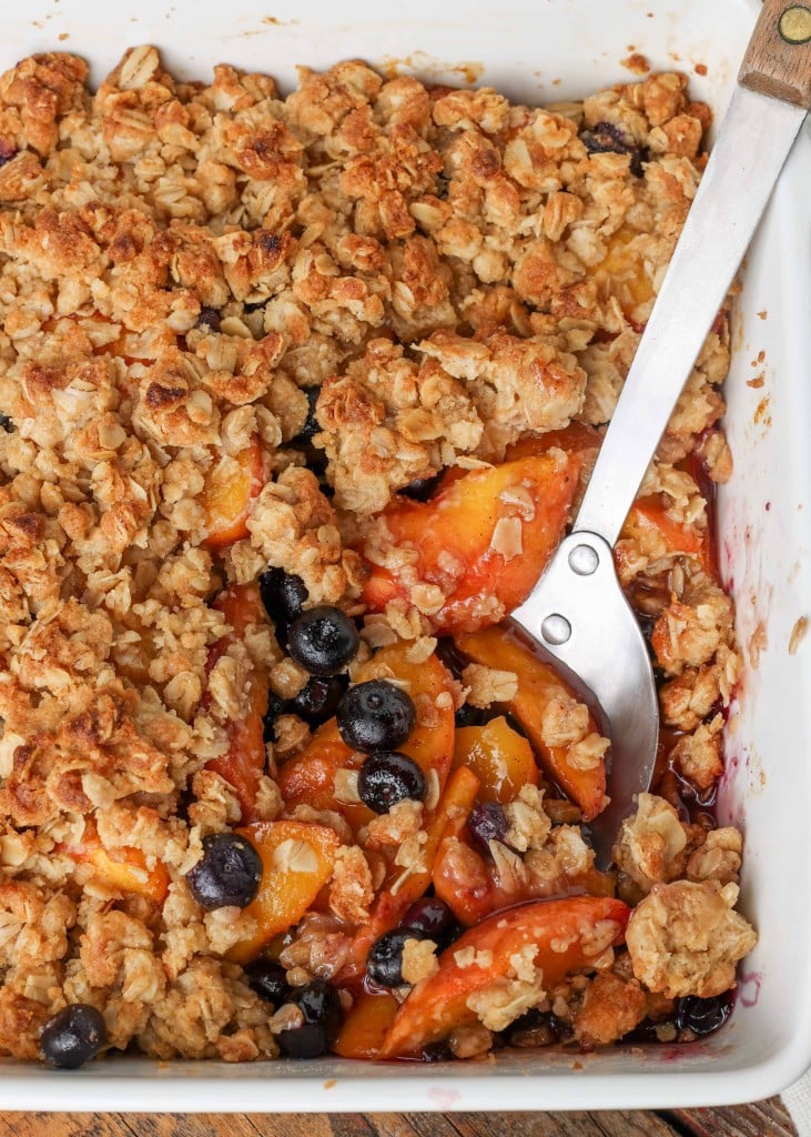 sliced peaches and blueberries are visible beneath the oat crust of the crisp in this top down shot of the pan with a metal spoon in it.
