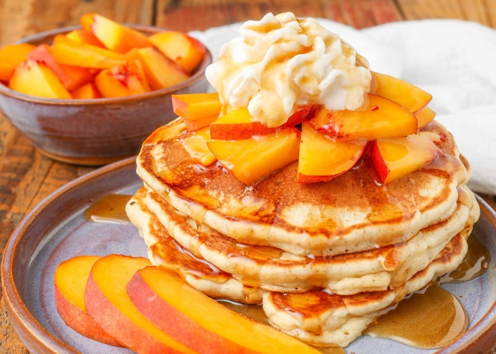 A horizontally aligned photo of a stack of pancakes on a light blue plate has been topped with whipped cream and pieces of fresh peach and drizzled with syrup.