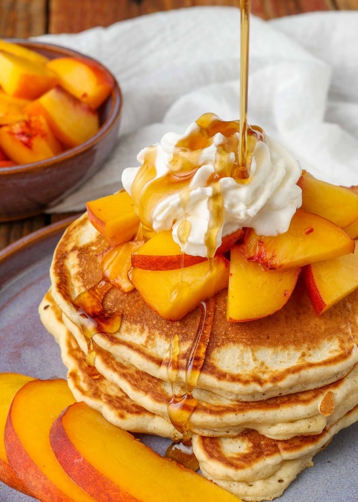 an action shot of pouring syrup over the stack of pancakes with peaches and whipped cream toppping.