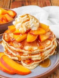 a vertically aligned photo of a stack of golden brown peach pancakes with pieces of peach as a topping with whipped cream and syrup.