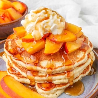 a vertically aligned photo of a stack of golden brown peach pancakes with pieces of peach as a topping with whipped cream and syrup.