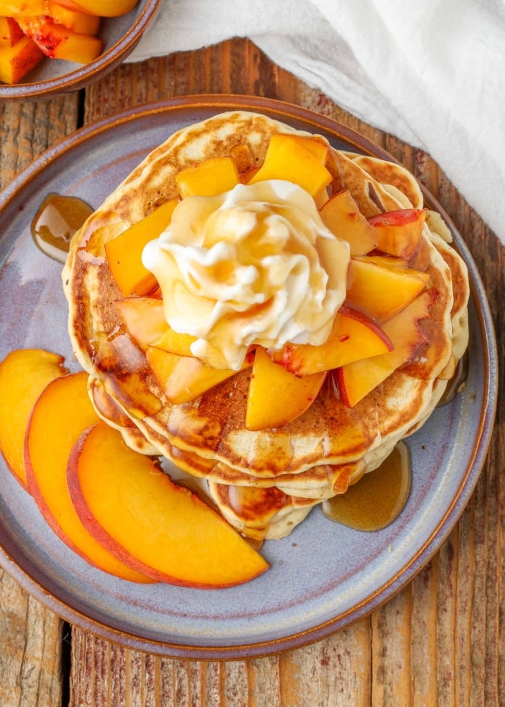 a vertically aligned, top down photo of a stack of golden brown peach pancakes with pieces of peach as a topping with whipped cream and syrup.