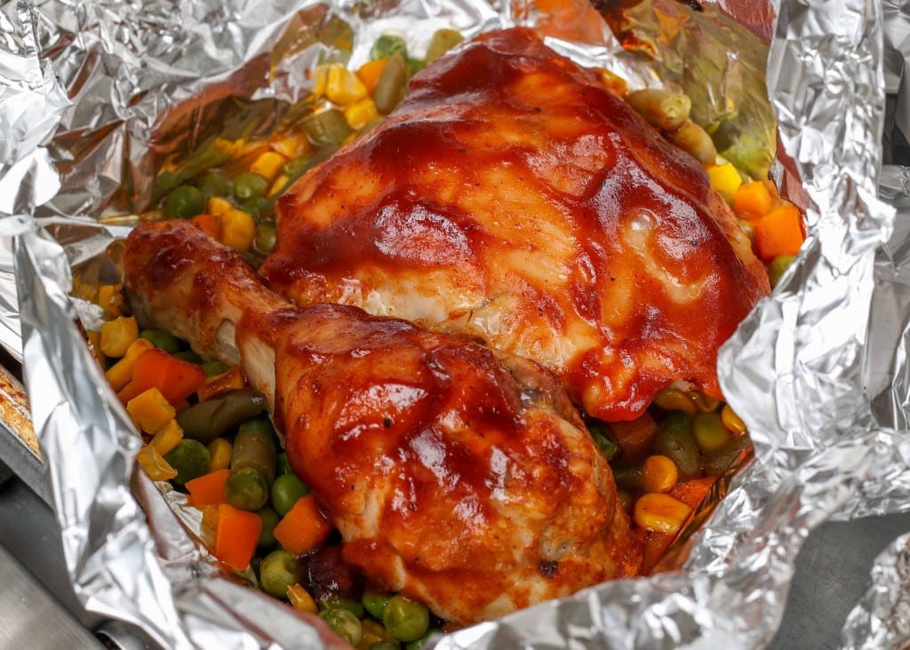 Overhead vertical shot of chicken brushed with BBQ sauce, served in a foil packet on a bed of peas, corn, and diced carrots