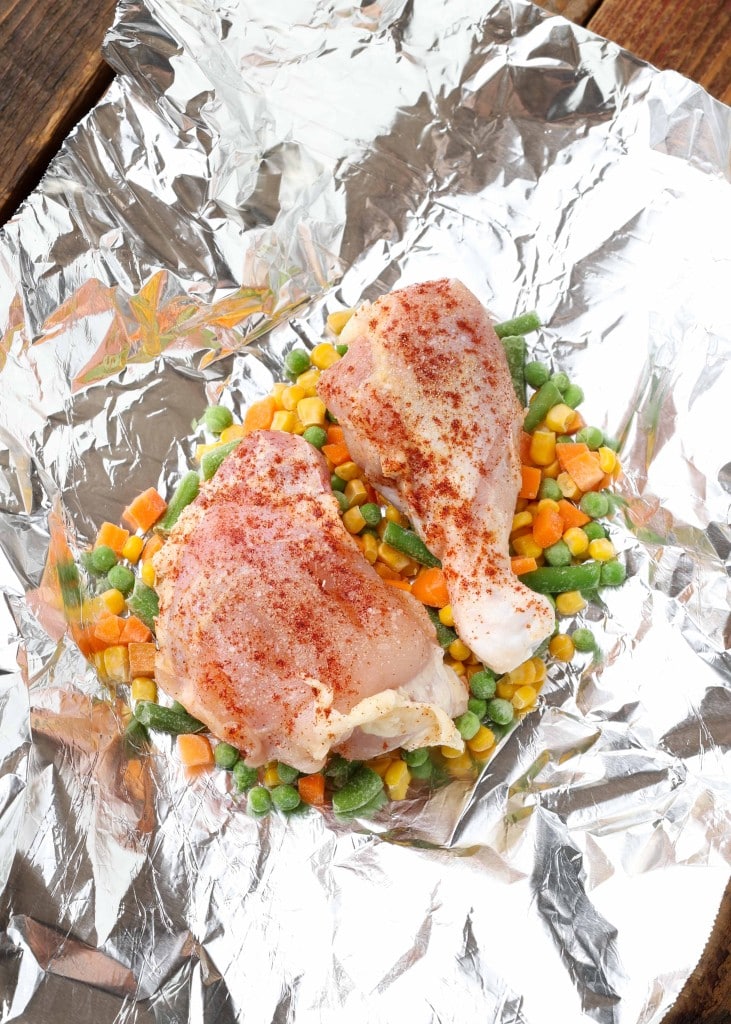 Overhead vertical shot of uncooked chicken in a foil packet on a bed of peas, corn, and diced carrots