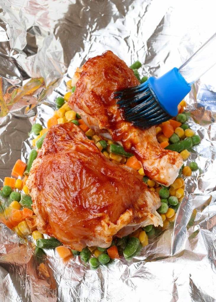 Overhead vertical shot of chicken brushed with BBQ sauce, served in a foil packet on a bed of peas, corn, and diced carrots