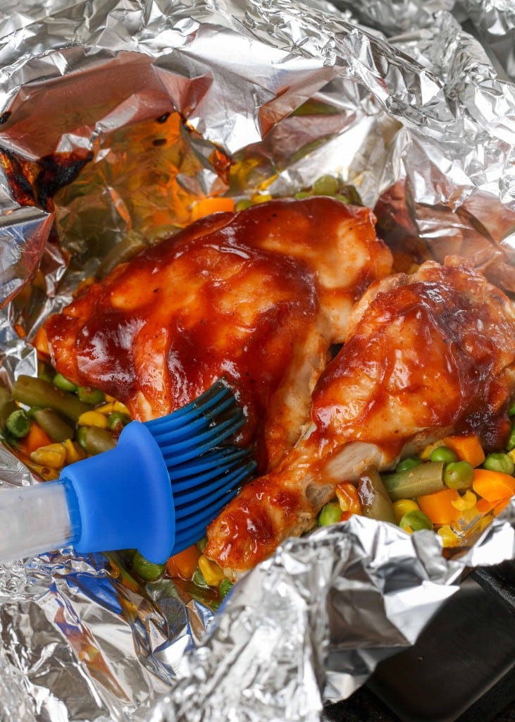 Overhead shot of chicken being brushed with BBQ sauce, served in a foil packet on a bed of peas, corn, and diced carrots
