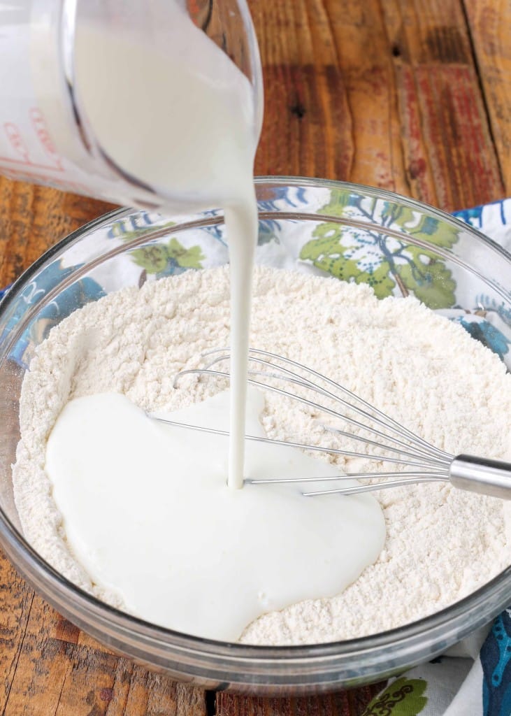 Overhead shot of flour and buttermilk mid-pour in a glass bowl with a silver whisk