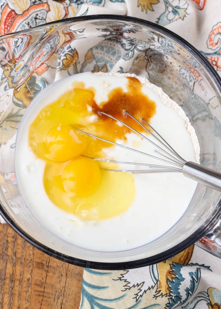 Overhead vertical shot of milk, flour, and eggs in a glass dish with a silver whisk