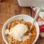 A vertically aligned image of a bowl of pumpkin crisp topped with vanilla ice cream, with a metal handled spoon sticking out.