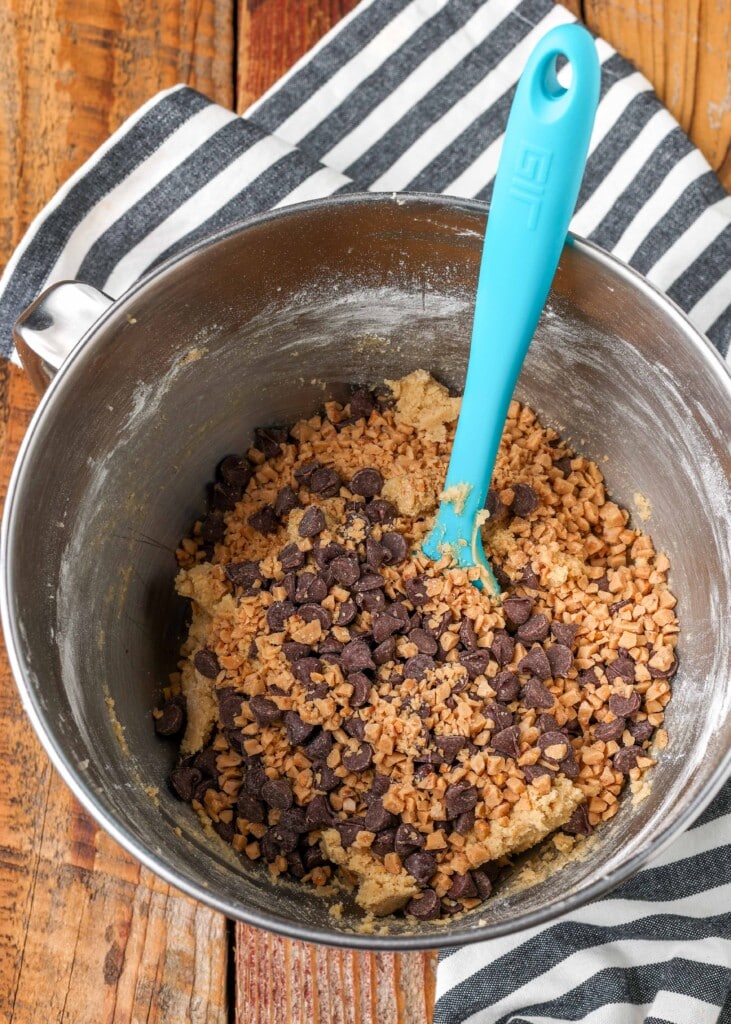 Chopped toffee and chocolate chips in a silver bowl with a blue spatula