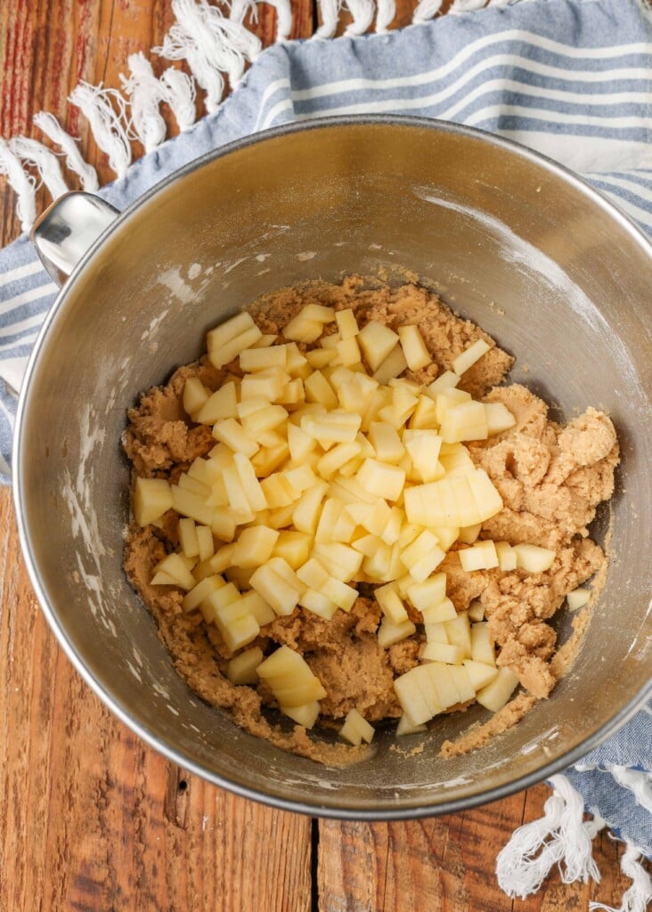 A metal mixing bowl filled with the ingredients for the apple cookies, with apple chunks piled high on top.
