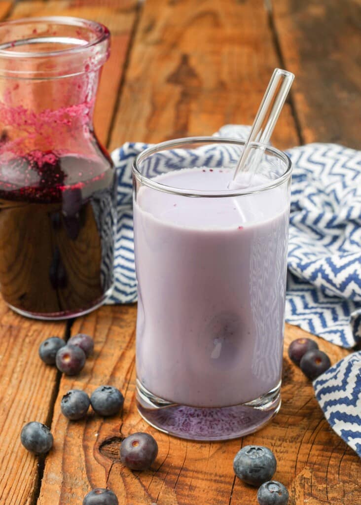 A vertical shot of the blueberry milk in a glass with a glass straw.