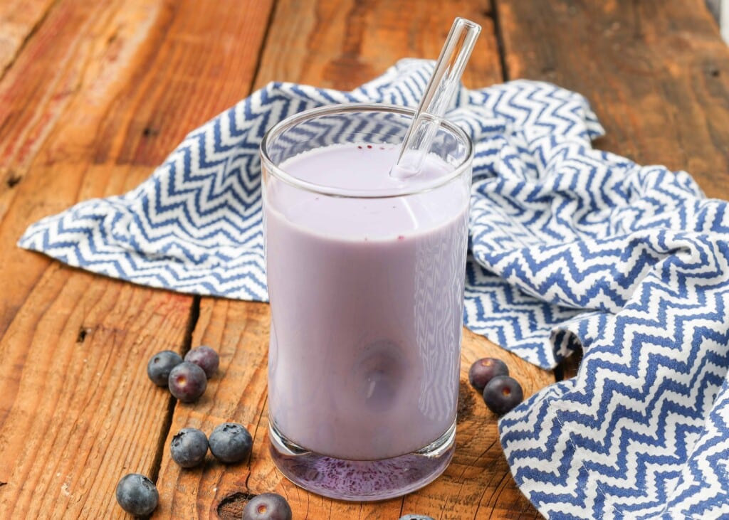 a horizontally aligned photograph of the drink on a wooden tabletop with blueberries scattered artfully around it and a tea towel visible in the background.