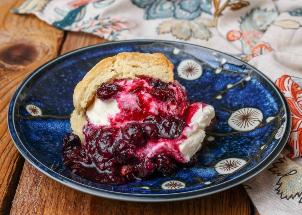a horizontal shot of blueberry shortcake with vanilla ice cream and blueberry compote on a dark blue ceramic plate.