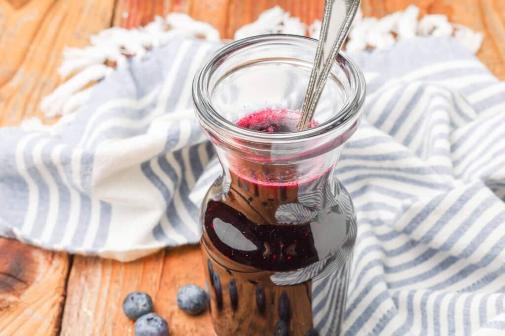 a horizontally aligned, close up shot of a clear glass carafe of blueberry simple syrup with a tea towel visible in the backgorund and blueberries scattered across the wooden tabletop.