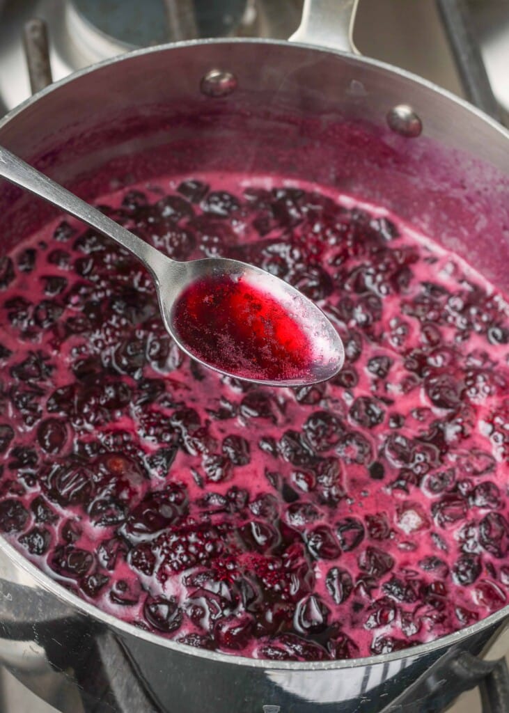 a spoon shows the color of the liquid above the pot of simmering blueberry mixture.