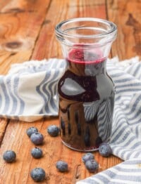 a vertically aligned shot of a clear glass carafe of blueberry simple syrup with a tea towel visible in the backgorund and blueberries scattered across the wooden tabletop.