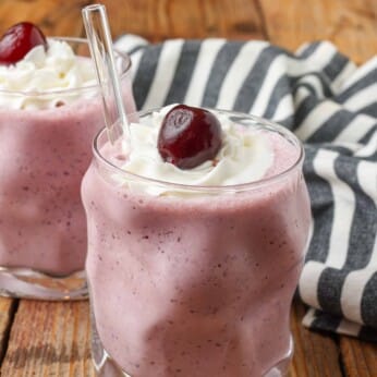 cherry milkshakes in glasses with glass straws topped with cherries and whipped cream