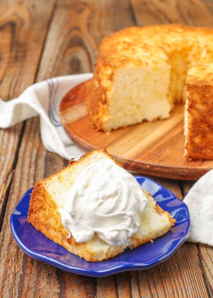 pineapple angel food cake on a cutting board with a whipped cream topped slice on a blue plate on a wooden table