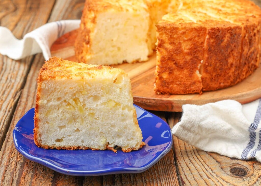 pineapple angel food cake on a cutting board with a whipped cream topped slice on a blue plate on a wooden table