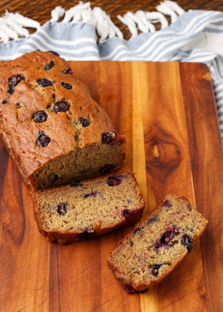 a loaf of blueberry banana bread on a wooden cutting board with a striped tea towel