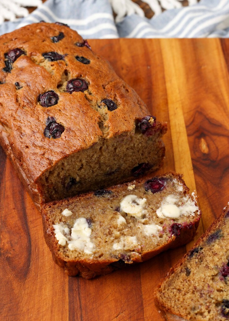 a loaf of banana blueberry bread on a cutting board with a buttered slice