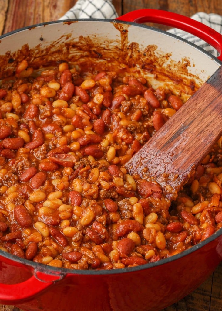 Baked beans with ground beef and bacon in red dutch oven with wooden spoon