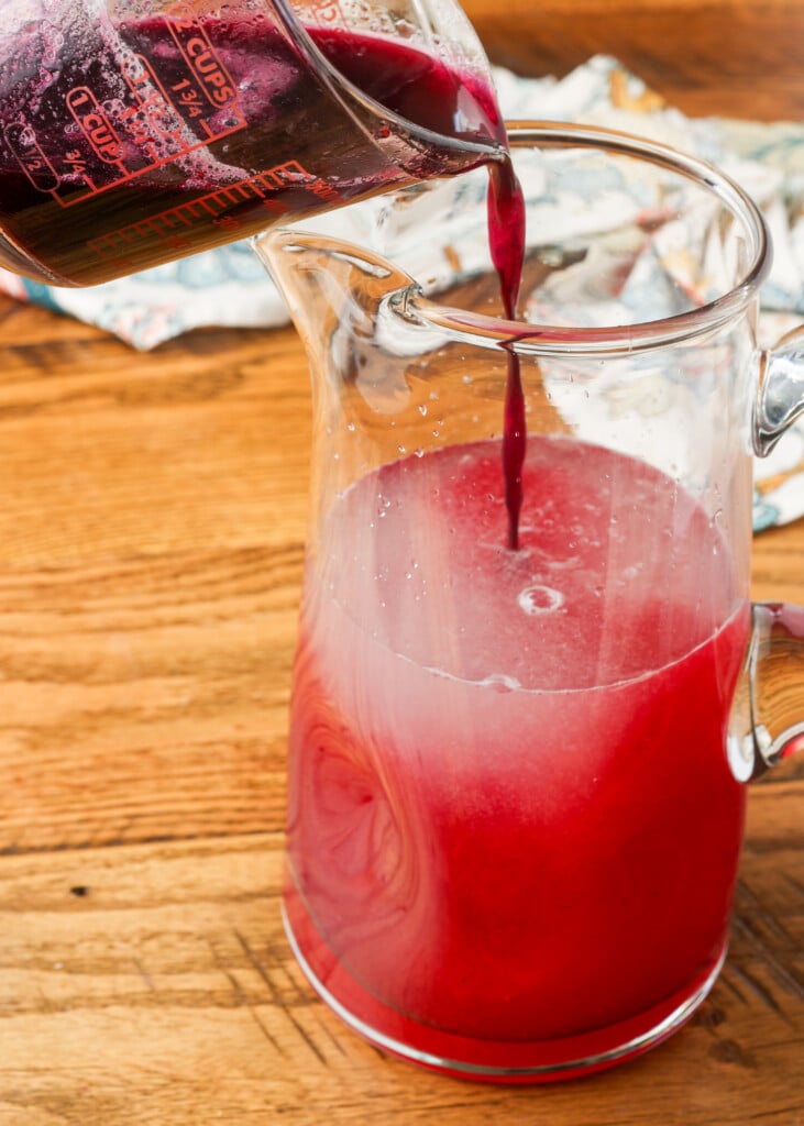 blueberry syrup being added to lemonade in a pitcher