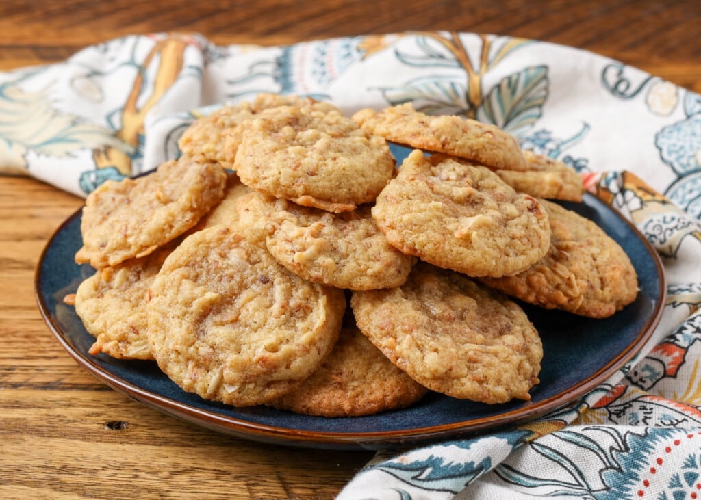 Chewy coconut cookies are piled high on a black ceramic plate over a wooden tabletop.