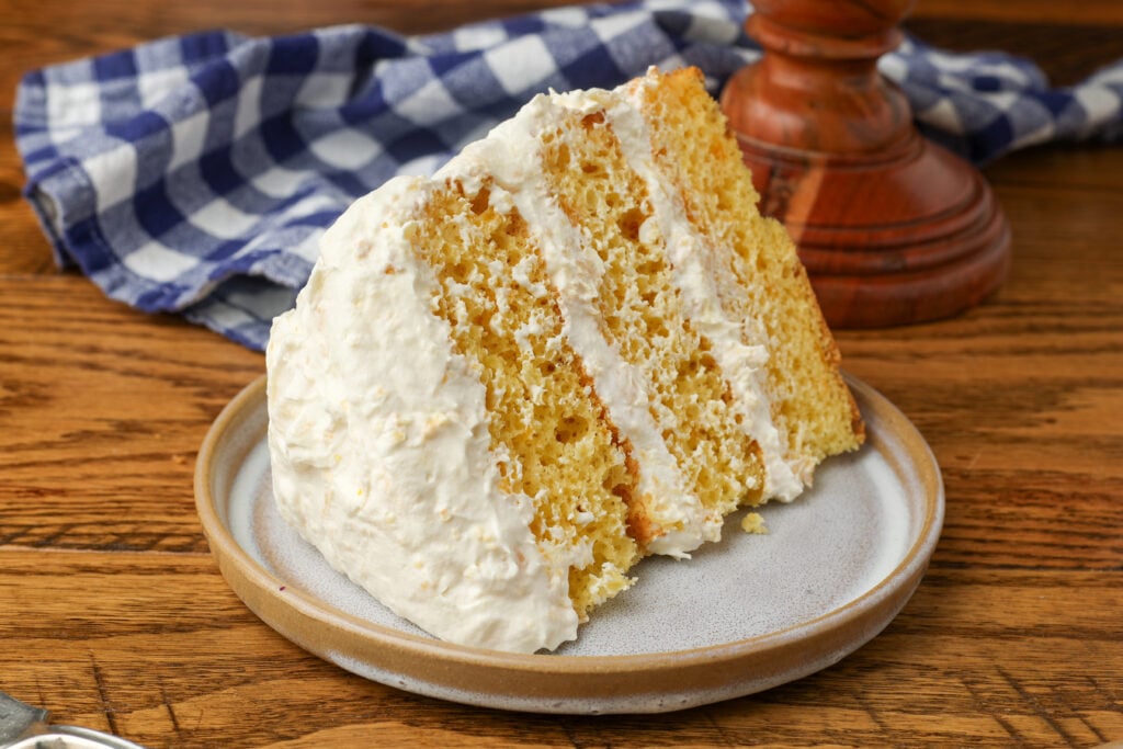 A wedge of pig pickin cake is laid on a small ceramic plate on a wooden tabletop with a blue and white checkered tea towel visible in the background.