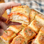 A vertically aligned close up image of a hand holding one of the chicken parm sliders in the foreground with the rest of the sliders visible in the background.