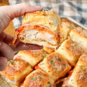 A vertically aligned close up image of a hand holding one of the chicken parm sliders in the foreground with the rest of the sliders visible in the background.