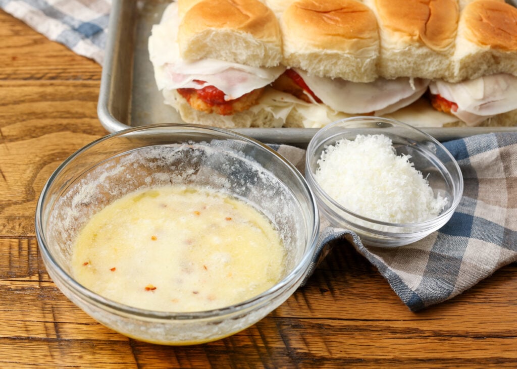A horizontally aligned image of the melted butter mixture, ready to be brushed onto the tops of the rolls with a smaller bowl of shredded parmesan nearby.