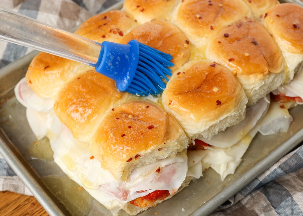 A horizontally aligned image of an action shot, showing the butter being brushed over the tops of the hawaiian rolls.