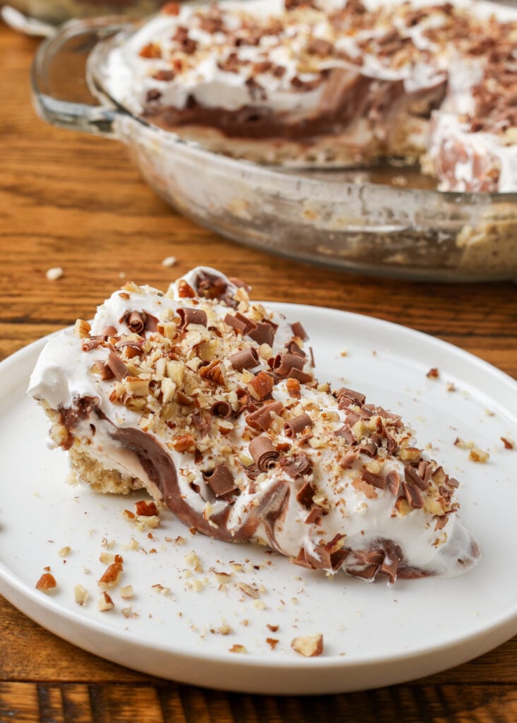 A vertically aligned image of a slice of possum pie on a white plate on a wooden tabletop with the rest of the pie visible in a dish in the background.