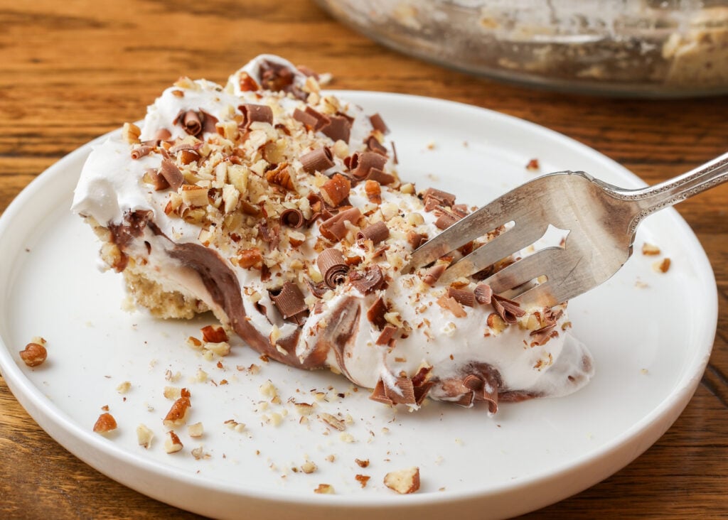 A horizontally aligned image of a slice of possum pie on a white ceramic plate with a fork reaching in to life a bite off of the end.